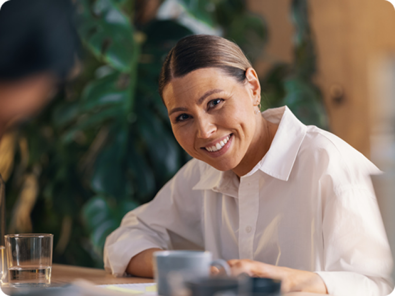 Dame som sit i eit kontorlandskap og jobbar, smiler og ser inn i kameraet